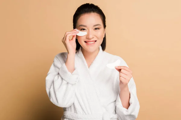 Feliz hermosa mujer asiática en albornoz aplicando parches en la cara sobre fondo beige - foto de stock