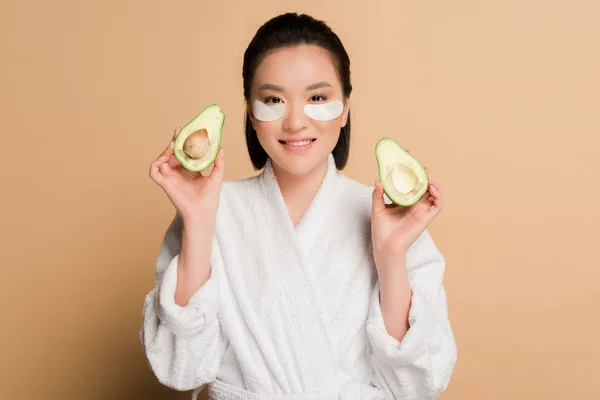 Happy beautiful asian woman in bathrobe with eye patches on face holding avocado halves on beige background — Stock Photo