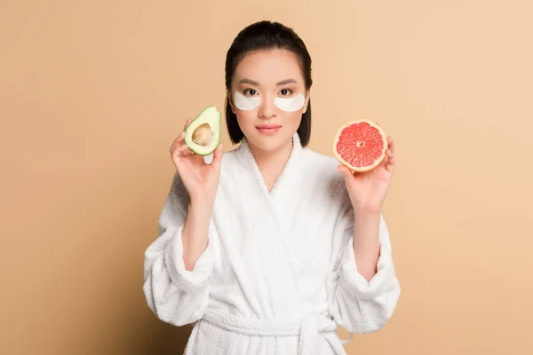 Beautiful asian woman in bathrobe with eye patches on face holding grapefruit and avocado halves on beige background — Stock Photo