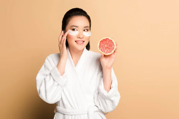 Sonriente hermosa mujer asiática en albornoz con parches en la cara sosteniendo la mitad de pomelo sobre fondo beige — Stock Photo