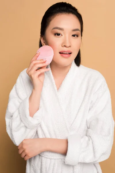 Красива азіатська жінка в халаті використовує щітку для очищення обличчя на бежевому фоні — стокове фото