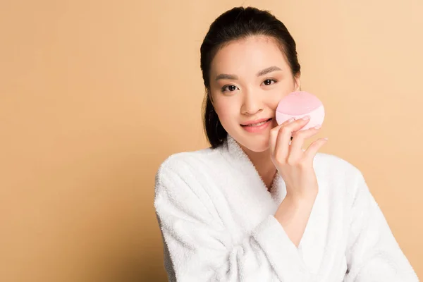 Beautiful asian woman in bathrobe using facial cleansing brush on beige background — Stock Photo