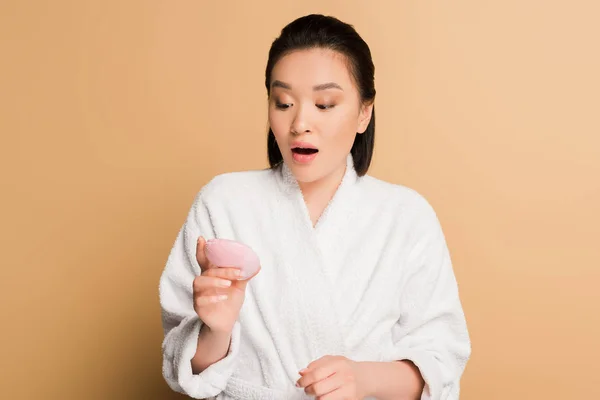 Schockiert schöne asiatische Frau im Bademantel mit Blick auf Gesichtsreinigungsbürste auf beigem Hintergrund — Stockfoto