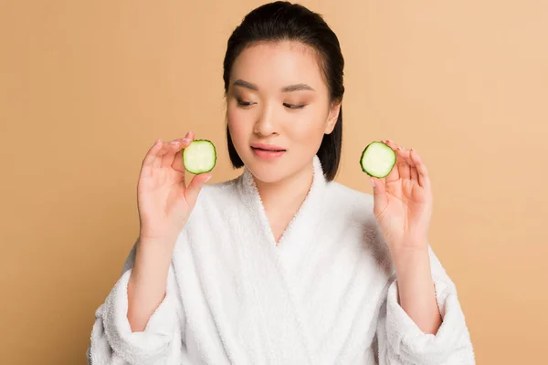 Beautiful asian woman in bathrobe with cucumber slices on beige background — Stock Photo
