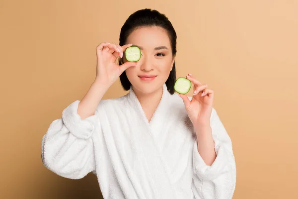 Lächelnd schöne asiatische Frau im Bademantel mit Gurkenscheiben auf beigem Hintergrund — Stockfoto