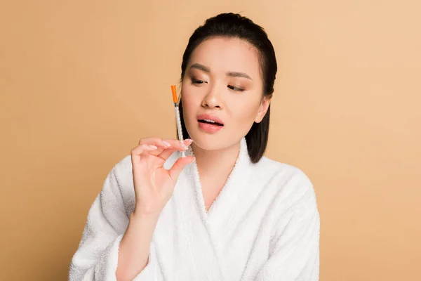 Hermosa mujer asiática en albornoz mirando jeringa para inyección de belleza sobre fondo beige - foto de stock