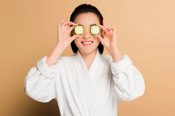 Sonriente hermosa mujer asiática en albornoz con rodajas de pepino en los ojos sobre fondo beige - foto de stock