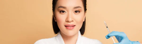 Hermosa mujer asiática en albornoz cerca de mano cosmetóloga con jeringa para inyección de belleza sobre fondo beige, tiro panorámico - foto de stock