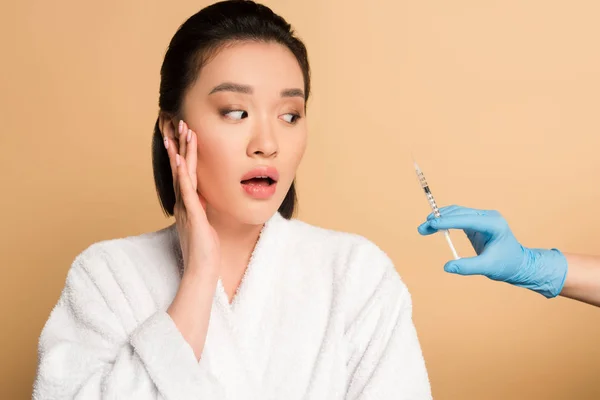 Peur belle asiatique femme en peignoir près de cosmétologue main avec seringue pour beauté injection sur fond beige — Photo de stock