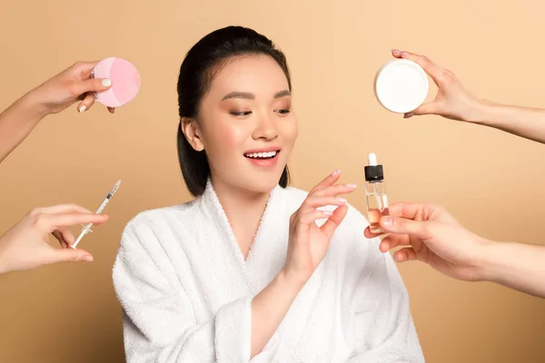 Sourire belle asiatique femme en peignoir près des mains avec brosse de nettoyage du visage, seringue, crème cosmétique et huile sur fond beige — Photo de stock