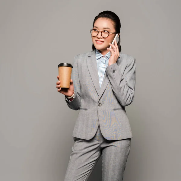 Sorridente jovem empresária em óculos segurando copo de papel e falando no smartphone em fundo cinza — Fotografia de Stock