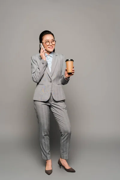 Smiling young businesswoman in eyeglasses holding paper cup and talking on smartphone on grey background — Stock Photo