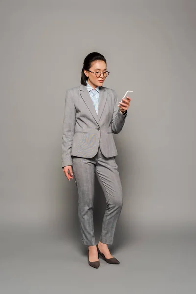 Young businesswoman in eyeglasses using smartphone on grey background — Stock Photo