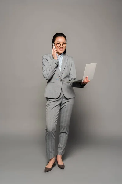 Усміхнена молода бізнес-леді в окулярах використовує ноутбук і розмовляє на смартфоні на сірому фоні — стокове фото