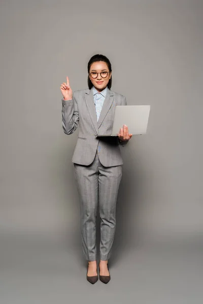 Усміхнена молода бізнес-леді в окулярах використовує ноутбук і показує жест ідеї на сірому фоні — стокове фото