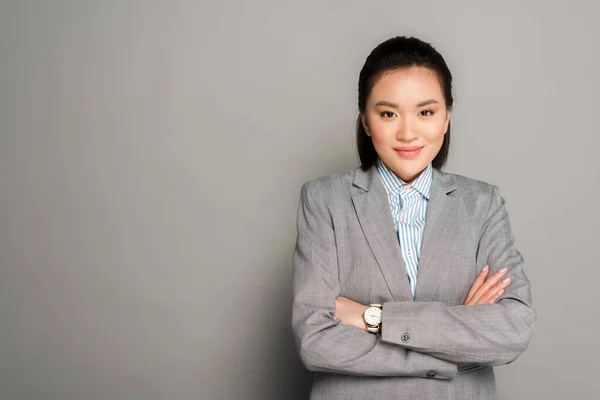 Jeune femme d'affaires souriante en costume avec les bras croisés sur fond gris — Photo de stock