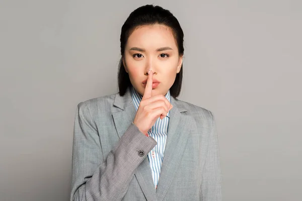 Junge Geschäftsfrau im Anzug zeigt shh-Geste auf grauem Hintergrund — Stockfoto