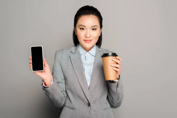 Щаслива молода бізнес-леді з паперовою чашкою тримає смартфон на сірому фоні — стокове фото