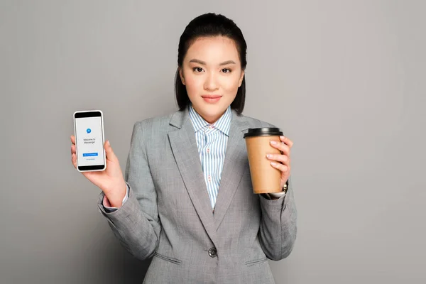 KYIV, UCRANIA - 11 de febrero de 2019: joven empresaria feliz con taza de papel que sostiene el teléfono inteligente con aplicación de mensajero sobre fondo gris - foto de stock