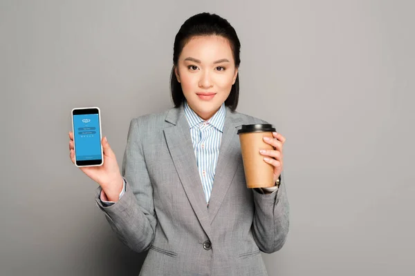 KYIV, UCRAINA - 11 FEBBRAIO 2019: felice giovane donna d'affari con coppa di carta che tiene smartphone con app skype su sfondo grigio — Foto stock