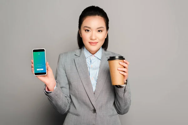 KYIV, UKRAINE - 11 FÉVRIER 2019 : heureuse jeune femme d'affaires avec tasse en papier tenant smartphone avec application twitter sur fond gris — Photo de stock