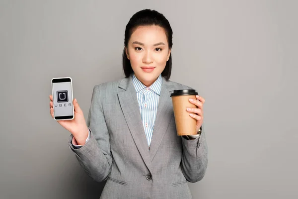 KYIV, UKRAINE - FEVEREIRO 11, 2019: jovem empresária feliz com copo de papel segurando smartphone com aplicativo uber no fundo cinza — Fotografia de Stock