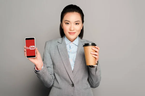 KYIV, UKRAINE - FEVEREIRO 11, 2019: jovem empresária feliz com copo de papel segurando smartphone com aplicativo youtube no fundo cinza — Fotografia de Stock
