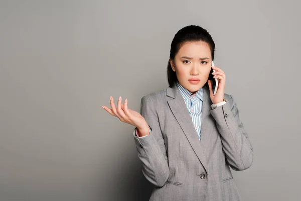 Запутавшаяся молодая деловая женщина в костюме разговаривает по смартфону на сером фоне — стоковое фото
