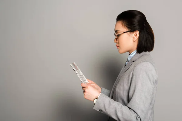 Seitenansicht der jungen Geschäftsfrau mit Brille beim Zeitungslesen auf grauem Hintergrund — Stockfoto