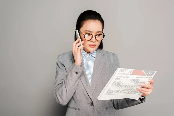 Joven mujer de negocios en gafas leyendo el periódico y hablando en el teléfono inteligente sobre fondo gris - foto de stock