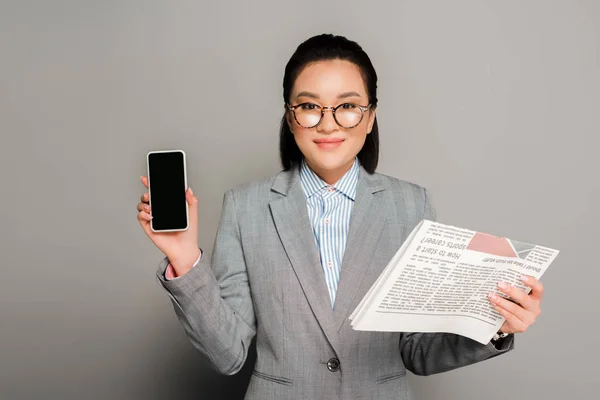 Junge Geschäftsfrau mit Brille hält Zeitung und Smartphone auf grauem Hintergrund — Stockfoto