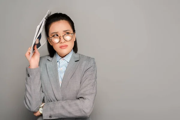 Traurige junge Geschäftsfrau mit Brille hält Zeitung auf grauem Hintergrund — Stockfoto