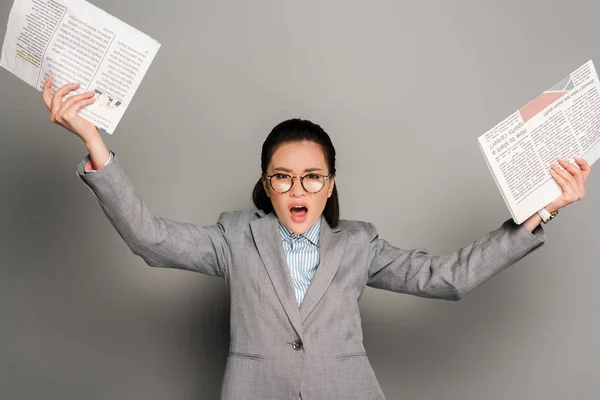 Angry young businesswoman in eyeglasses holding newspapers on grey background — Stock Photo