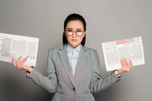 Sad young businesswoman in eyeglasses holding newspapers on grey background — Stock Photo