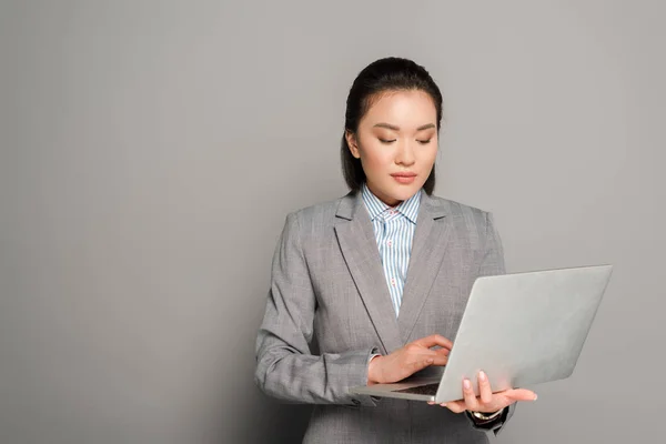 Jovem empresária de terno usando laptop no fundo cinza — Fotografia de Stock