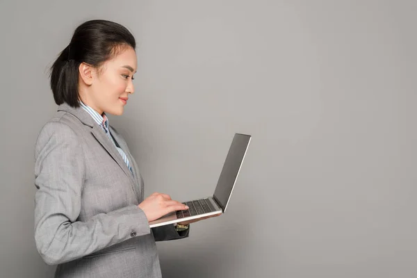 Vista lateral de la joven mujer de negocios sonriente en traje usando el ordenador portátil sobre fondo gris - foto de stock