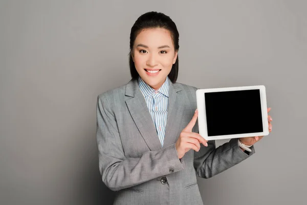 Feliz joven empresaria de traje sosteniendo tableta digital sobre fondo gris - foto de stock