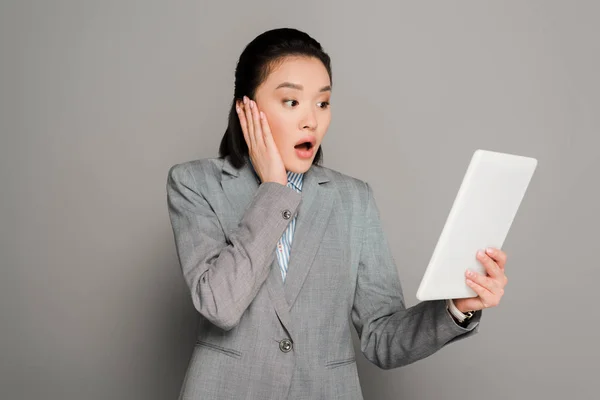 Shocked young businesswoman in suit using digital tablet on grey background — Stock Photo