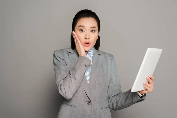 Sorprendido joven mujer de negocios en traje usando tableta digital sobre fondo gris - foto de stock