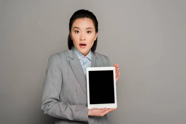 Шокированная молодая бизнесвумен в костюме держит цифровой планшет на сером фоне — стоковое фото