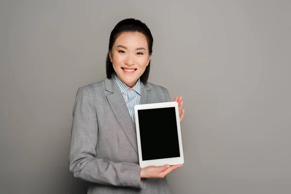 Улыбающаяся молодая деловая женщина в костюме держит цифровой планшет на сером фоне — стоковое фото