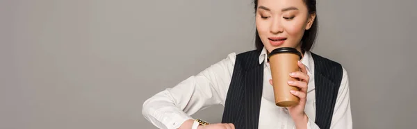 Улыбающаяся молодая деловая женщина смотрит на наручные часы и пьет кофе изолированный на сером, панорамный снимок — стоковое фото