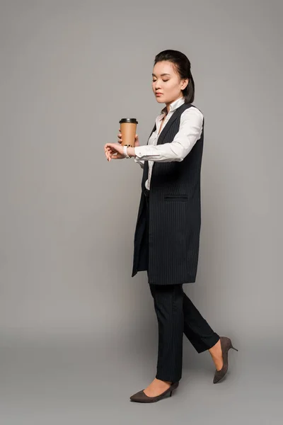 Young businesswoman looking at wristwatch and drinking coffee on grey background — Stock Photo