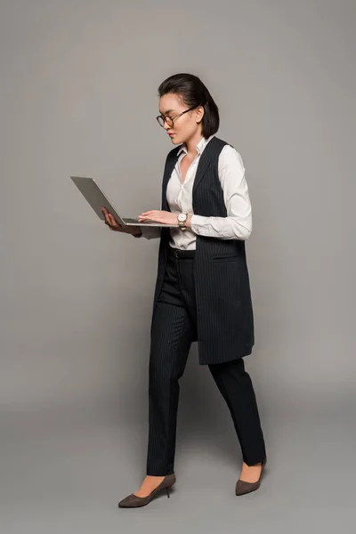 Jovem empresária em óculos usando laptop em fundo cinza — Fotografia de Stock
