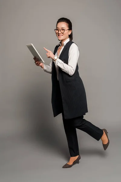 Glückliche junge Geschäftsfrau mit Brille zeigt mit dem Finger auf digitales Tablet auf grauem Hintergrund — Stockfoto