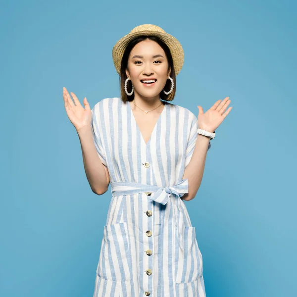 Sorrindo morena asiática menina em vestido listrado e chapéu de palha com braços abertos isolados em azul — Fotografia de Stock