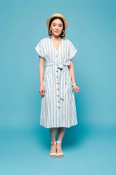 Brunette asian girl in striped dress and straw hat on blue background — Stock Photo