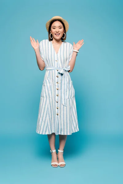 Smiling brunette asian girl in striped dress and straw hat with open arms on blue background — Stock Photo