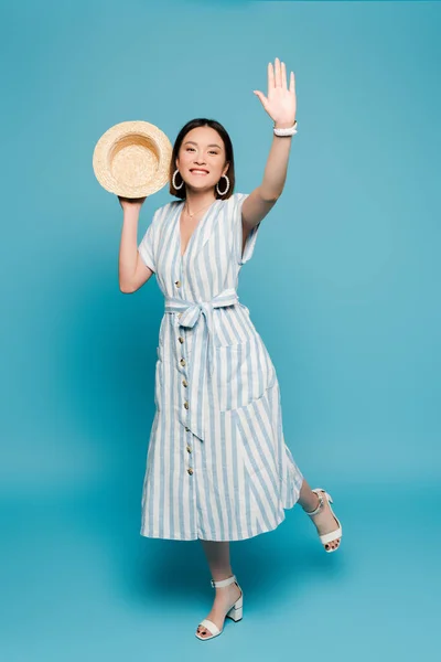 Smiling brunette asian girl in striped dress and straw hat waving hand on blue background — Stock Photo