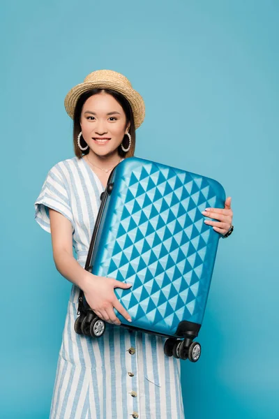 Усміхнена брюнетка азіатська дівчина в смугастій сукні і солом'яному капелюсі з дорожньою сумкою на синьому фоні — стокове фото
